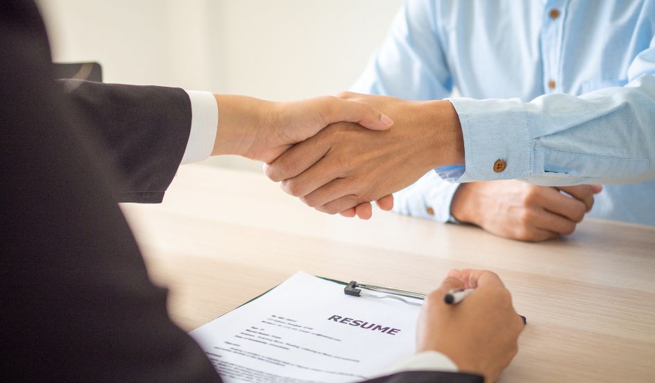 two men shaking hands at an interview with a clipboard with a resume on it