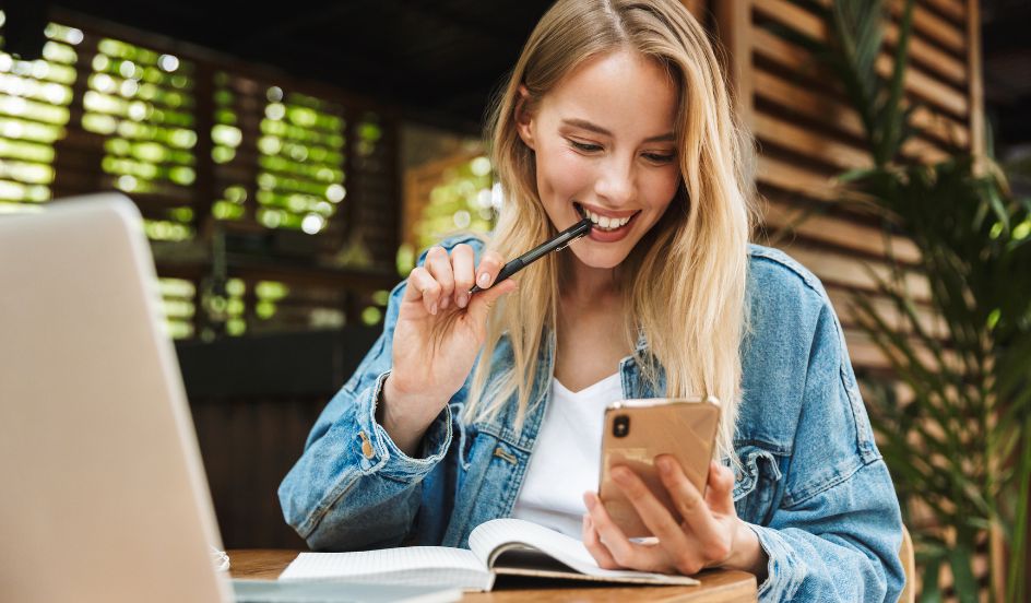 woman on smartphone smiling while in front of laptop and notebook