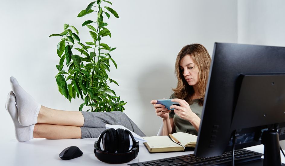 woman procrastinating sitting at a desk on her phone with her feet up