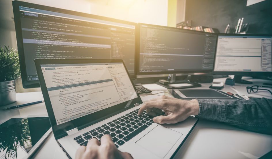 hands on laptop keyboard with coding on screen and computers in background