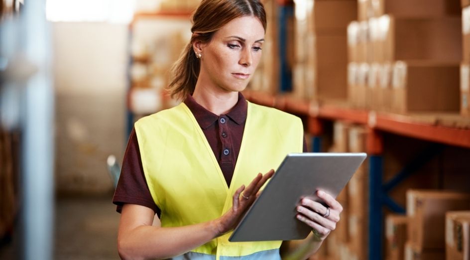 a woman in a yellow vest holds a tablet and stands in front of rows of boxes