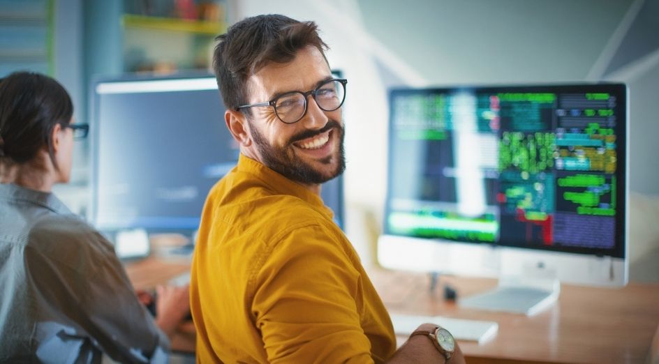 a man in a yellow shirt and glasses, smiling in front of a computer in an office