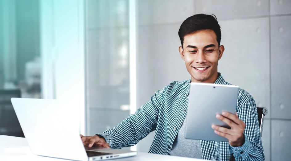 smiling man sits at his desk, looking at paper with his hand on laptop