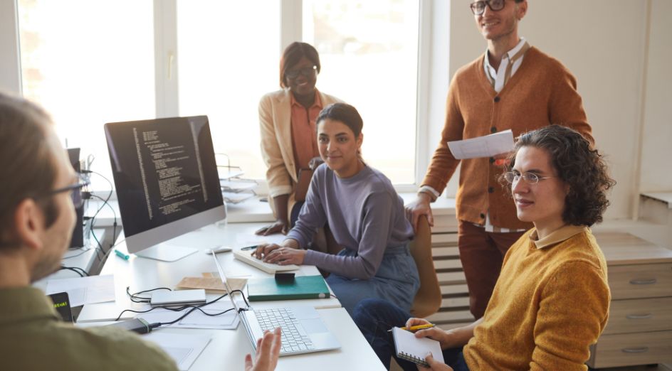 a group of people sit and stand around a large desk, discussing code on a computer screen